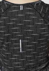 Svart tränings topp med ett prickigt mönster med vita fläckar, rund halsringning och dragkedjedetalj i ryggen. Tillverkad av fukttransporterande material.