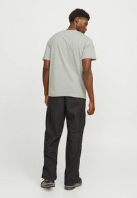 Mann in hellgrauem T-Shirt, schwarzen Cargo-Hosen und Sneakers, der steht und über seine Schulter blickt, vor einem schlichten weißen Hintergrund.