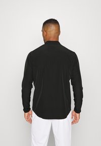 Black long-sleeve shirt with a smooth texture, collar, and rounded hem. The fabric appears lightweight and fitted. Worn with white pants.