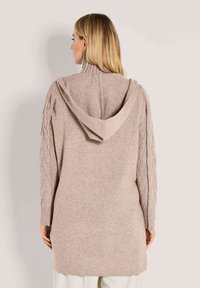 Frau mit blonden Haaren trägt einen langen beigefarbenen Strickpullover mit Kapuze und strukturierten Ärmeln, steht mit dem Rücken zu einer schlichten Hintergrund.