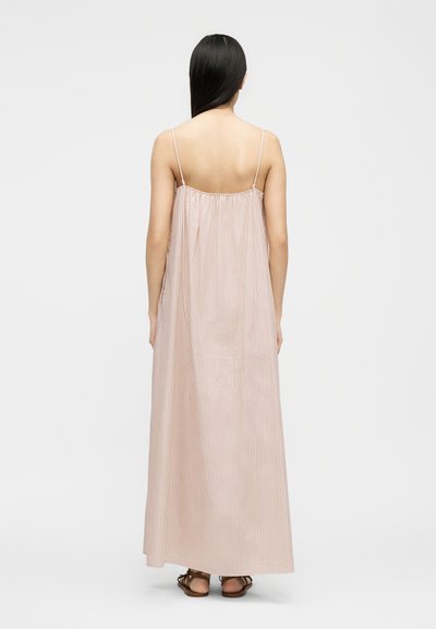 Γυναίκα με μακριά μαύρα μαλλιά, φορόντας ένα αμάνικο, μακρύ ως το πάτωμα φόρεμα με ανοιχτό ροζ και λευκές ρίγες και καφέ σανδάλια, στέκεται βλέποντας προς τα πίσω.