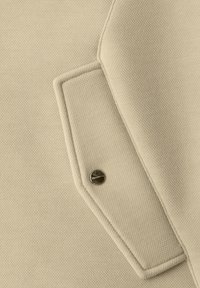 Beiges, strukturiertes Gewebe mit einer Lasche, die durch einen Metall-Druckknopf gesichert ist, mit detaillierten Nähten entlang der Kanten.