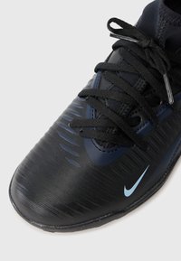 Čierna atletická obuv značky Nike s textúrovaným dizajnom, zaoblenou špičkou, modrými akcentmi a čiernymi šnúrkami na svetlosivom pozadí.