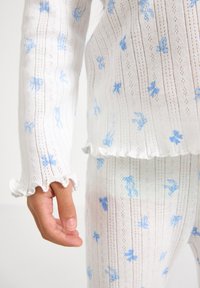 Ensemble de détente en tricot blanc avec des motifs de nœuds bleus. Présente des ourlets ondulés et un tissu texturé avec de petits trous. Manches avec des poignets volantés.