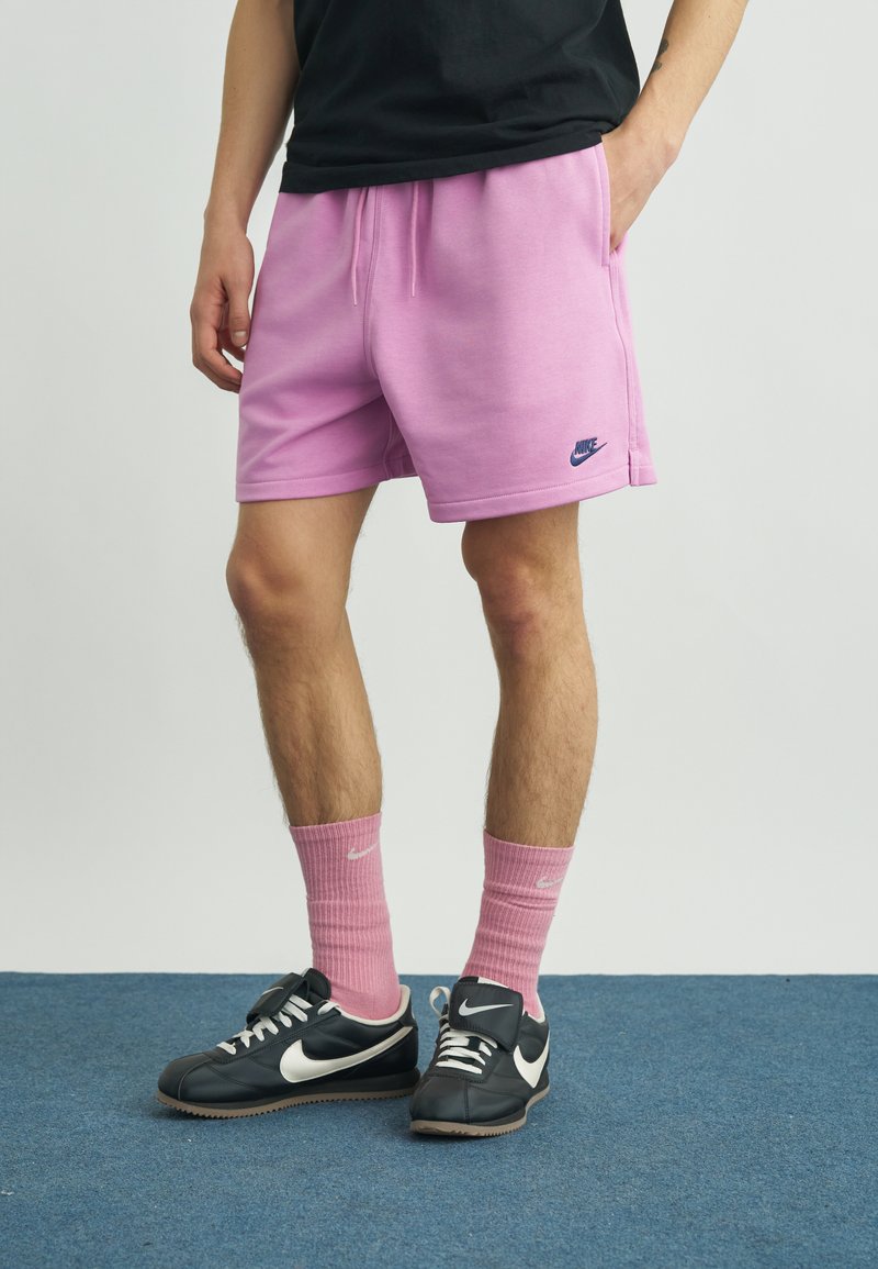 Άνδρας που φοράει ροζ σορτς Nike, ροζ κάλτσες που ταιριάζουν και μαύρα παπούτσια Nike, στέκεται σε μπλε χαλί.