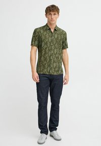 Camicia verde a maniche corte con una stampa di foglie, abbinata a jeans scuri e sneaker grigio chiaro. La camicia presenta un design con bottoni.