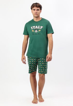 Hombre vistiendo una camiseta de manga corta temática de Italia en color verde con Mickey Mouse y rebanadas de pizza, pantalones cortos a juego con el mismo patrón, descalzo sobre fondo blanco.
