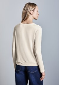 Helles beiges Langarmshirt mit einem runden Ausschnitt und glattem Stoff, kombiniert mit dunkelblauen Jeans. Verfügt über eine lockere Passform und einen lässigen Saum.