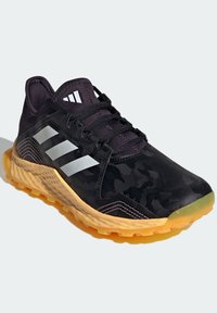 Czarne buty sportowe z kamuflażem, trzy białe paski po bokach, teksturowana gumowa podeszwa, pomarańczowe akcenty i czarne sznurowadła.