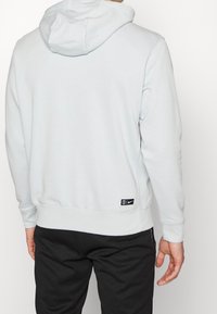 Sudadera azul claro con capucha ajustable, dobladillo y puños acanalados. Presenta una etiqueta negra con el logo en la parte inferior izquierda. Combinada con pantalones oscuros.