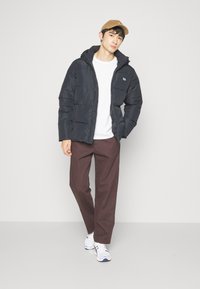 Doudoune noire avec capuche, fermeture éclair et logo sur la poitrine, associée à un t-shirt blanc, un pantalon marron et des baskets blanches.
