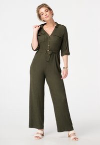 Combinaison texturée vert olive avec deux poches poitrine, fermeture à boutons sur le devant, taille ceinturée et design à jambes larges. Associée à des sandales rose clair.
