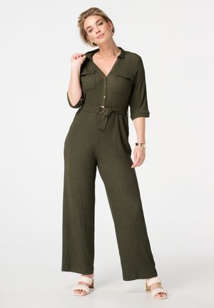 Combinaison texturée vert olive avec deux poches poitrine, fermeture à boutons sur le devant, taille ceinturée et design à jambes larges. Associée à des sandales rose clair.