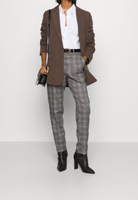 Blazer marrone sopra una camicia bianca, pantaloni grigi a quadri sartoriali e tacchi a punta neri. Borsa nera con una tracolla lucida.