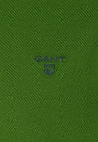 Tissu vert texturé avec le nom de marque GANT brodé en bleu marine et un logo en forme de bouclier centré.