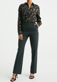 Kvinna i mörkgröna högmidjade raka byxor, en blommig långärmad skjorta instoppad, och svarta öppna högklackade sandaler.