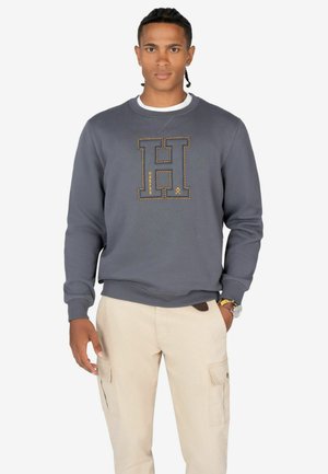 Sudadera gris con un gran logo de "H" bordado en amarillo, con puños y dobladillo acanalados. Combinada con pantalones cargo beige.