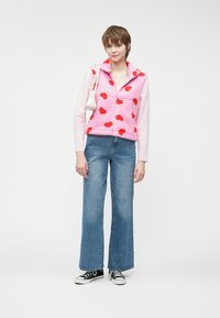 Ροζ γούνινο γιλέκο με κόκκινες καρδιές, συνδυασμένο με ανοιχτό μπλε τζιν παντελόνι καμπάνα και μαύρα αθλητικά παπούτσια. Κρατάει μία μικρή λευκή τσάντα.