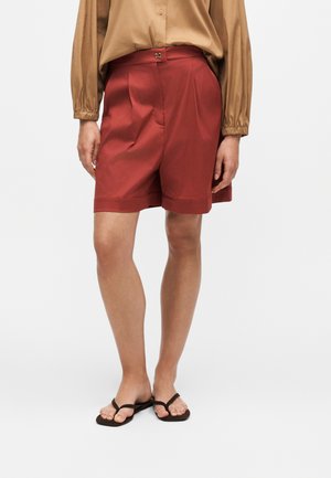Personne portant un short plissé couleur rouille avec une fermeture à bouton, une chemise beige à manches longues et des tongs noires, debout sur un fond blanc.