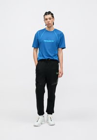 T-shirt atlético azul com o logótipo da Reebok, combinado com calças de fato pretas com detalhes refletivos e cintura elástica. Ténis brancos completam o visual.