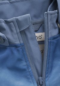 Blauwe stof met ritsluiting en platte kraag, met een gladde textuur en stikdetails. Label gedeeltelijk zichtbaar aan de binnenkant, met aanduiding van het merk.