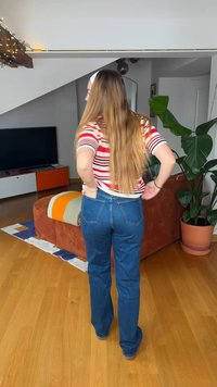 Γυναίκα με μακριά μαλλιά, που φοράει ριγέ πουκάμισο και μπλε τζιν, στέκεται μέσα σε σαλόνι με τηλεόραση, πορτοκαλί καναπέ και μεγάλο γλαστράκι.