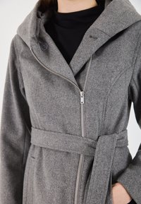 Cappotto di lana grigio con cintura, zip diagonale e ampio colletto, indossato sopra un top nero, mano parzialmente visibile nella tasca del cappotto.