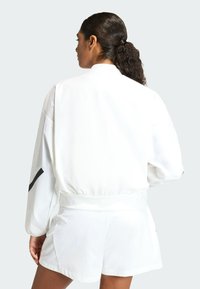 Bijela sportska jakna s elastičnim pojasom, dugim rukavima i crnim detaljima. Nošena uz odgovarajuće bijele kratke hlače, minimalnog dizajna.