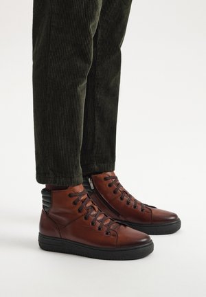 Brązowe skórzane sneakersy za kostkę z czarnymi podeszwami, z okrągłymi sznurówkami i żebrowanymi akcentami na pięcie, noszone z ciemnozielonymi spodniami z kordurowej tkaniny.