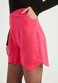 Klart rosa shorts med en slät yta, som har sidofickor och en scalloped nederkant. Stylat med svarta långärmade tröjor och genomskinliga strumpbyxor.