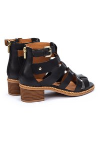 Sandalias de tacón de piel negra con tiras entrelazadas y cremalleras laterales. Cuentan con un tacón de madera apilada y un interior color tostado.