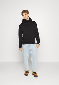 Sort hættebluse med forlomme og snor i hætten, parret med lyseblå jeans og sort-orange sportsko. Minimalistisk design og afslappet pasform.