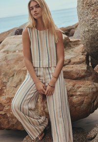 Young woman with long blonde hair wearing sleeveless striped top and wide-leg pants standing among large rocks by the sea.