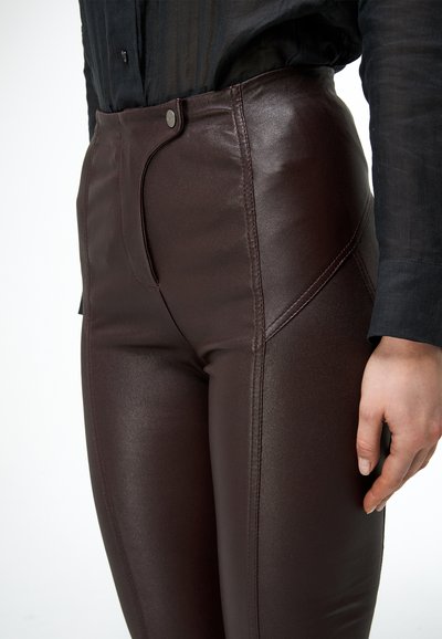 Zbliżenie osoby noszącej ciemnobrązowe skórzane spodnie z wysokim stanem z przodem zapinanym na guziki i detalami przeszyć, w połączeniu z czarną koszulą z długim rękawem.