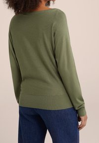 Pull vert à manches longues avec ourlet et poignets côtelés, fabriqué en tissu tricoté doux. Porté avec un jean en denim bleu, vue de dos.