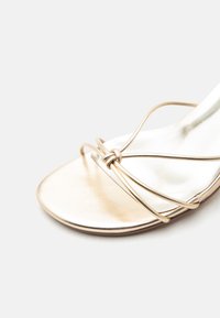 Sandale dorée à bout ouvert avec de fines lanières métalliques se croisant et formant un noeud à l'avant sur un fond blanc épuré.