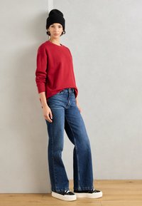 Rotes Sweatshirt mit langen Ärmeln, kombiniert mit blauen Jeans. Das Model trägt eine schwarze Beanie und schwarze Sneakers mit weißen Sohlen. Modischer Look.