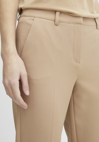 Beige op maat gemaakte broek met een gladde textuur, strakke lijnen, zijzakken en een slim fit, met een hand dichtbij de tailleband.
