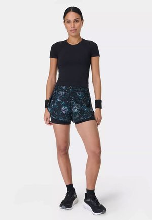 Vrouw staand, gekleed in een zwarte strakke T-shirt, sportkorte broek met patroon, zwarte polsbandjes, zwarte sokken en zwarte hardloopschoenen tegen een witte achtergrond.