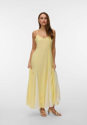 Kobieta w jasnożółtej sukience maxi bez rękawów na cienkich ramiączkach i beżowych sandałach na obcasie, stojąca na jednolitym białym tle.