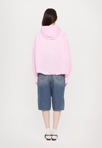 Rosa kariertes Kapuzenshirt mit langen Ärmeln, kombiniert mit knielangen, ausgewaschenen blauen Jeansshorts. Weiße Socken und schwarze Schuhe runden das Outfit ab.