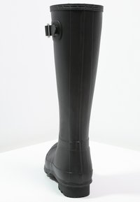 Botas de lluvia de goma negras hasta la rodilla con suela plana, detalle de hebilla en la parte trasera y diseño estriado en la parte superior. Textura exterior suave.