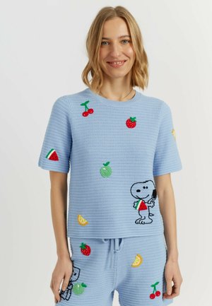 Donna sorridente che indossa un top azzurro chiaro lavorato a maglia e pantaloncini con toppe di frutta e Snoopy che tiene una fetta di anguria.