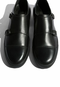Czarne skórzane mokasyny z dwiema regulowanymi sznurówkami, zaokrąglonym czubkiem i gładkim, błyszczącym wykończeniem; teksturowana gumowa podeszwa.