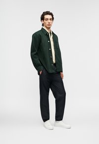 Πράσινο σακάκι από κορντόνι πάνω από μια κρεμ πουκάμισο, συνδυασμένο με μαύρο παντελόνι και λευκά αθλητικά παπούτσια· διαθέτει κουμπιά κλεισίματος και άνετη εφαρμογή.