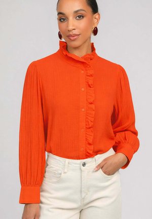 Femme portant un chemisier orange vif à manches longues avec un devant à volants et un col montant, associé à un pantalon blanc, posant avec une main dans la poche.