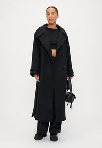 Melna vilnas maisījuma virsjakas ar plašu apkakli un dubultā pogojuma dizainu. Modelis tur mazu melnu somiņu. Pieguļošs melns tops un platai bikses.