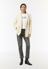Cremfarbener gerippter Blazer, weißes T-Shirt mit Grafik, graue gerade Jeans und schwarze Knöchelstiefeletten. Bemerkenswerte Details sind Doppelknöpfe und säumiger Saum.