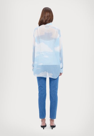 Chemise légère et transparente bleu clair avec un motif de nuages ; manches longues, ourlet arrondi, portée sur un jean skinny bleu et des talons noirs.