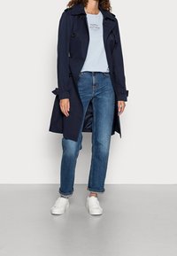 Abrigo de trench navy con cierre de botones y cintura ceñida, llevado sobre una camiseta azul clara y jeans azules, combinado con zapatillas blancas.